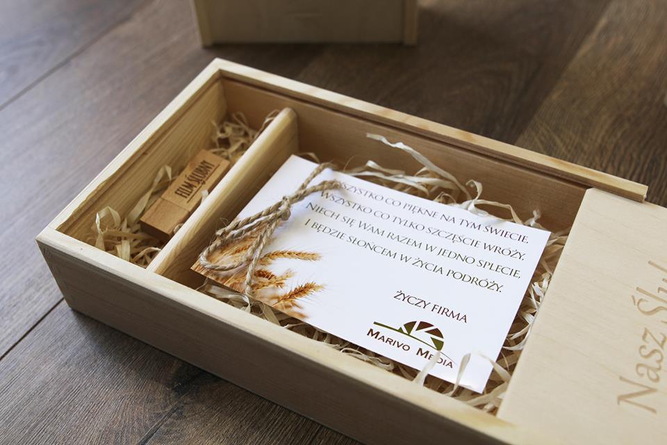 Drewniane pudełko z pendrivem 'Film Ślubny', kartką z życzeniami i logo Marivo Media, wypełnione dekoracyjną wełną drzewną, prezent na drewnianej podłodze.
