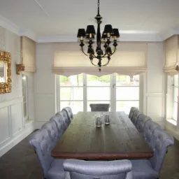 Elegancka jadalnia z długim drewnianym stołem, szarymi krzesłami z guzikami, beżowymi roletami w oknach i ozdobnym lustrem w złotej ramie na ścianie w kremowe pasy.