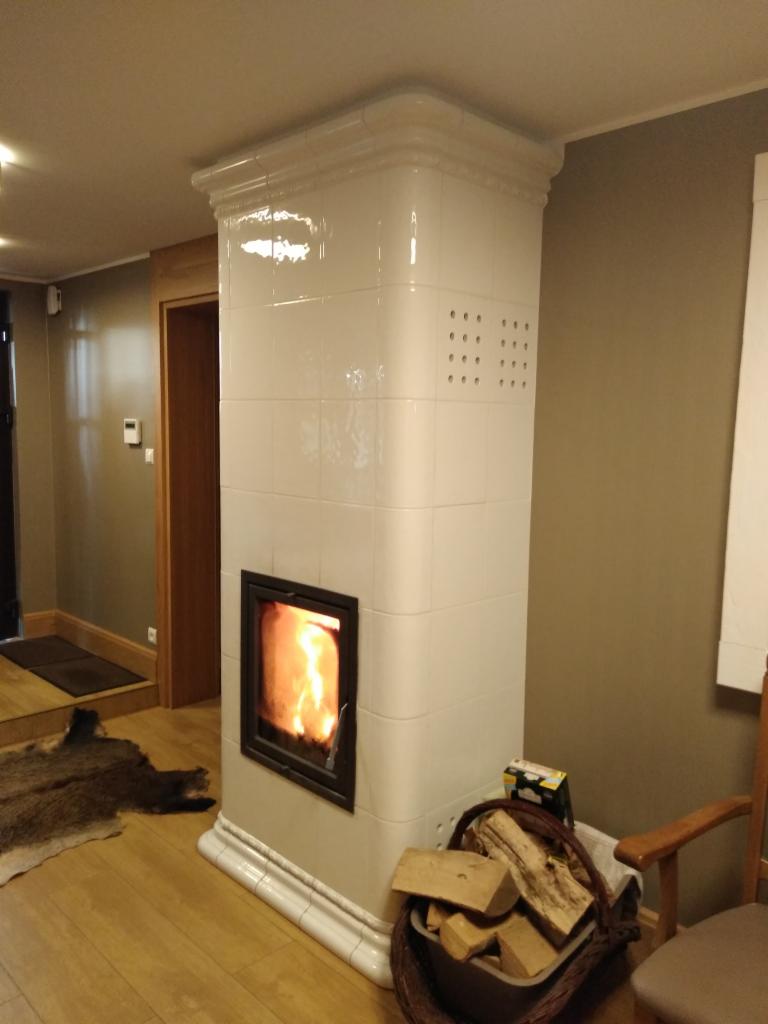 Biały, kaflowy piec kominkowy z paleniskiem, w tle drewniana podłoga i fragment skóry zwierzęcej, obok kosz z drewnem i krzesło.