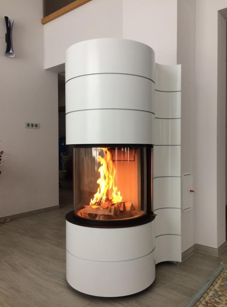 Nowoczesny, cylindryczny kominek z przeszklonym paleniskiem, płomienie tańczące wokół drewna, biała obudowa z poziomymi zdobieniami, jasne wnętrze z drewnianą podłogą.