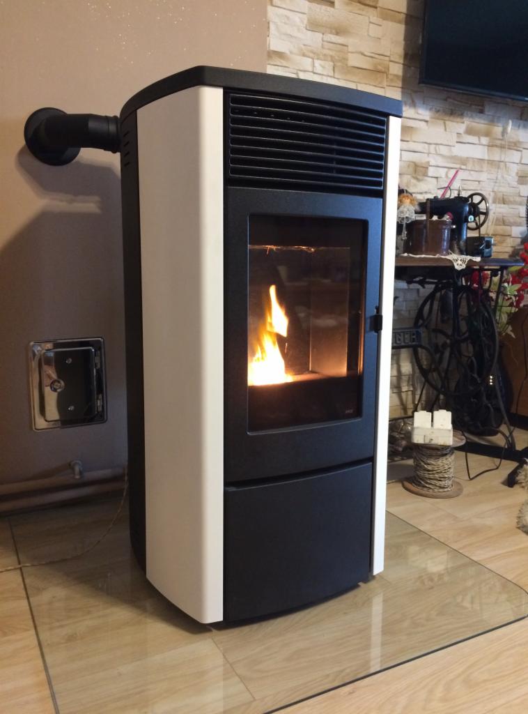 Nowoczesny piec na pellet z płomieniem widocznym przez szybę, umieszczony na szklanej płycie ochronnej, z widocznym przewodem kominowym po lewej stronie.