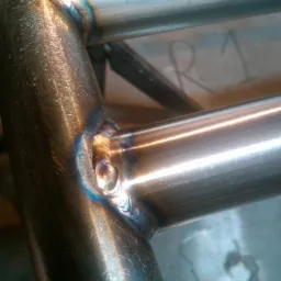 Szczegółowe zbliżenie na spawany narożnik ramy z rur stalowych, widoczne ślady spawania i opalizujące przebarwienia termiczne. W tle widoczny numer 'R 1' naniesiony markerem.