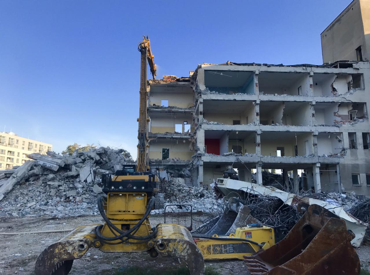 Żółta maszyna budowlana z długim ramieniem kruszy resztki budynku z odsłoniętymi piętrami, gruzowisko w tle, błękitne niebo.