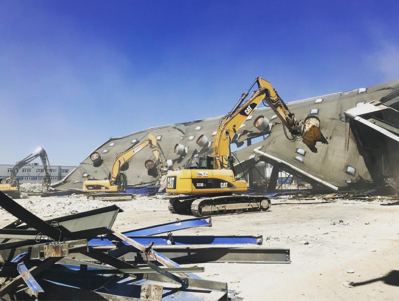 Żółte koparki CAT kruszą betonową konstrukcję budynku z okrągłymi otworami w dachu, na placu budowy w słoneczny dzień. W tle widoczny budynek biurowy.