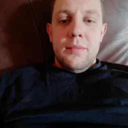 Portret mężczyzny w ciemnej koszulce na brązowym skórzanym fotelu, z lekkim zarostem i neutralnym wyrazem twarzy, oświetlony naturalnym światłem.