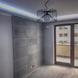 Pok&oacute;j z betonową ścianą z paneli, nowoczesna lampa wisząca, surowy beton na podłodze, widoczne gniazdka elektryczne, drzwi balkonowe z widokiem na inny budynek.