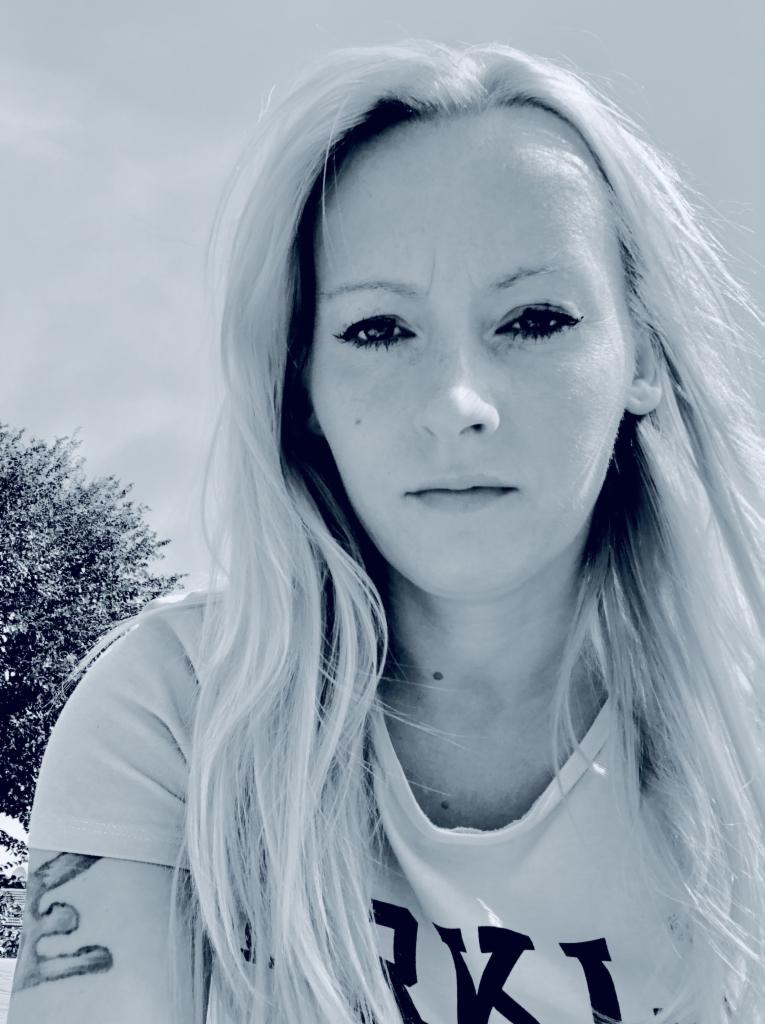 Portret kobiety z długimi blond włosami i tatuażem na ramieniu, na tle nieba i drzewa. Zdjęcie w odcieniach szarości, z wyrazistym makijażem oczu.