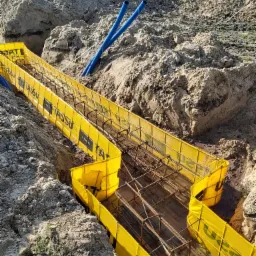 Wykonanie fundamentów: wykop z żółtą szalunką LÄMMI, wewnątrz zbrojenie stalowe, obok widoczne niebieskie rury i wykopana ziemia.