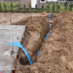 Wykop w ziemi z widocznymi niebieskimi rurami osłonowymi i rurą w kolorze terakoty, prawdopodobnie instalacja pod ogrzewanie podłogowe, widoczny betonowy fundament budynku, szpadel wbity w ziemię...