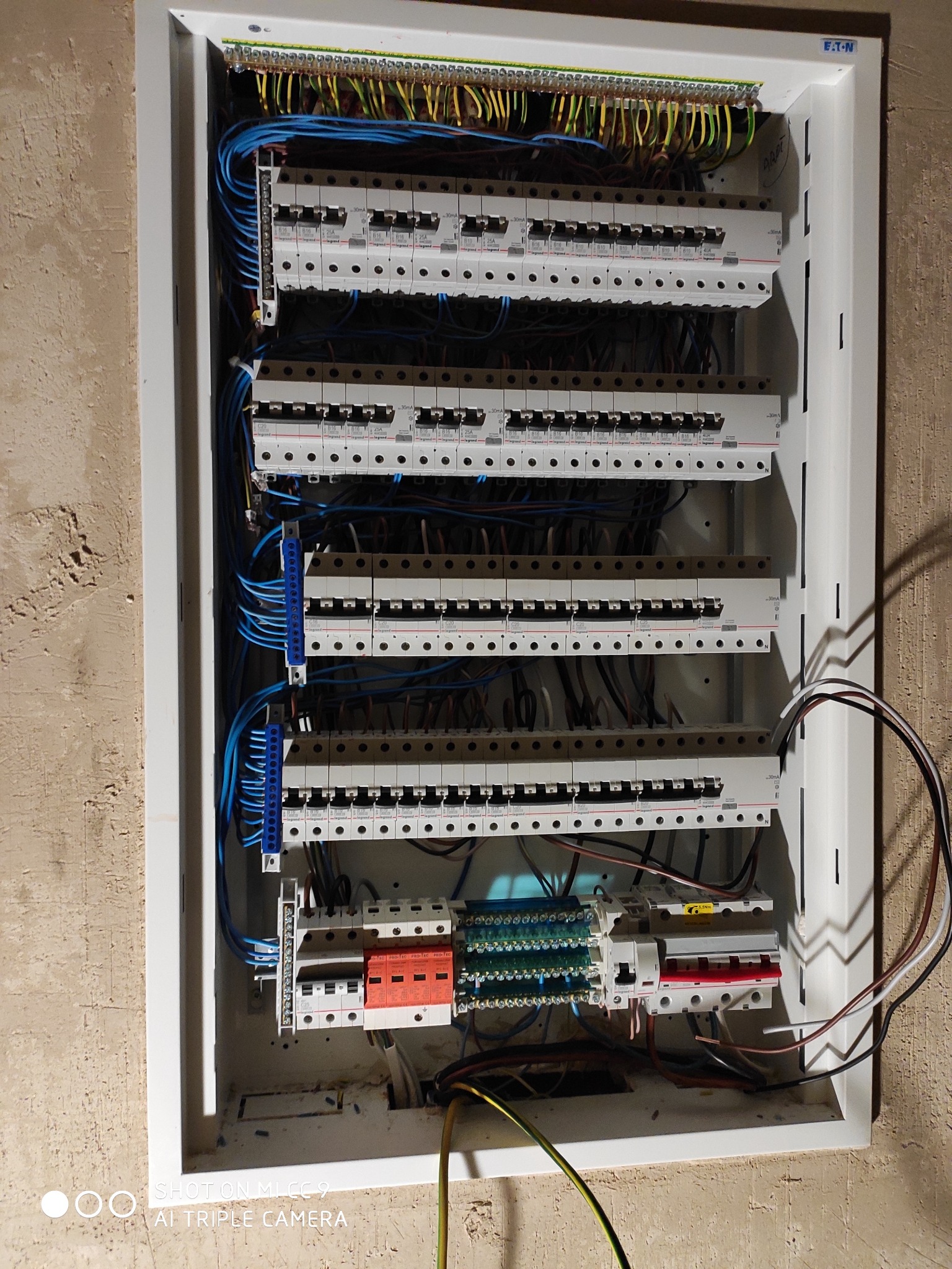 Otwarta skrzynka rozdzielcza z widocznymi bezpiecznikami, listwami zaciskowymi i kablami w różnych kolorach, umieszczona na nierównej ścianie z widoczną fakturą.