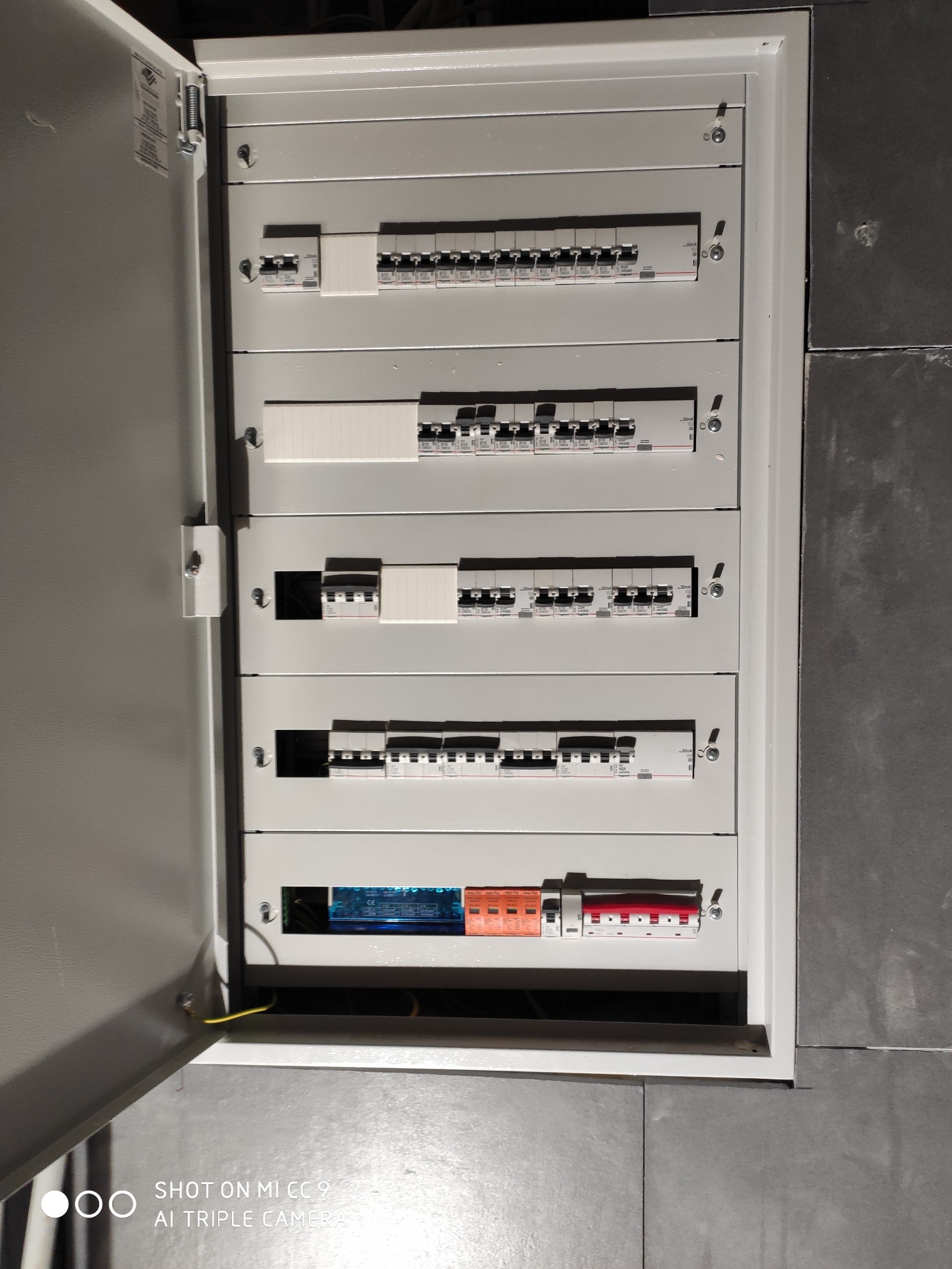 Otwarta skrzynka elektryczna z widocznymi bezpiecznikami i wyłącznikami różnicowoprądowymi, zainstalowana na szarej ścianie.
