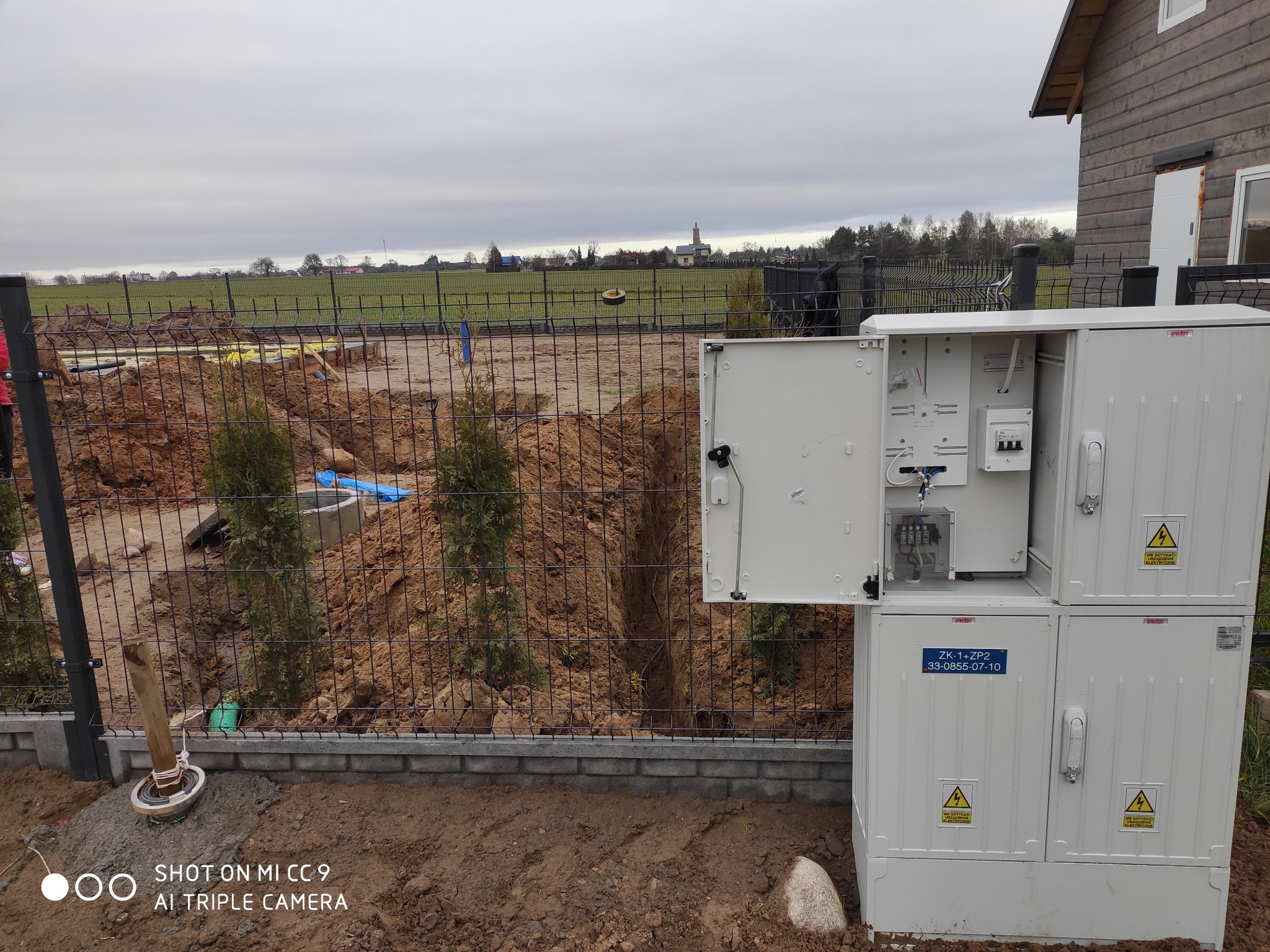 Biała skrzynka elektryczna z otwartymi drzwiczkami na tle wykopu, ogrodzenia i krajobrazu wiejskiego z kościołem w oddali.