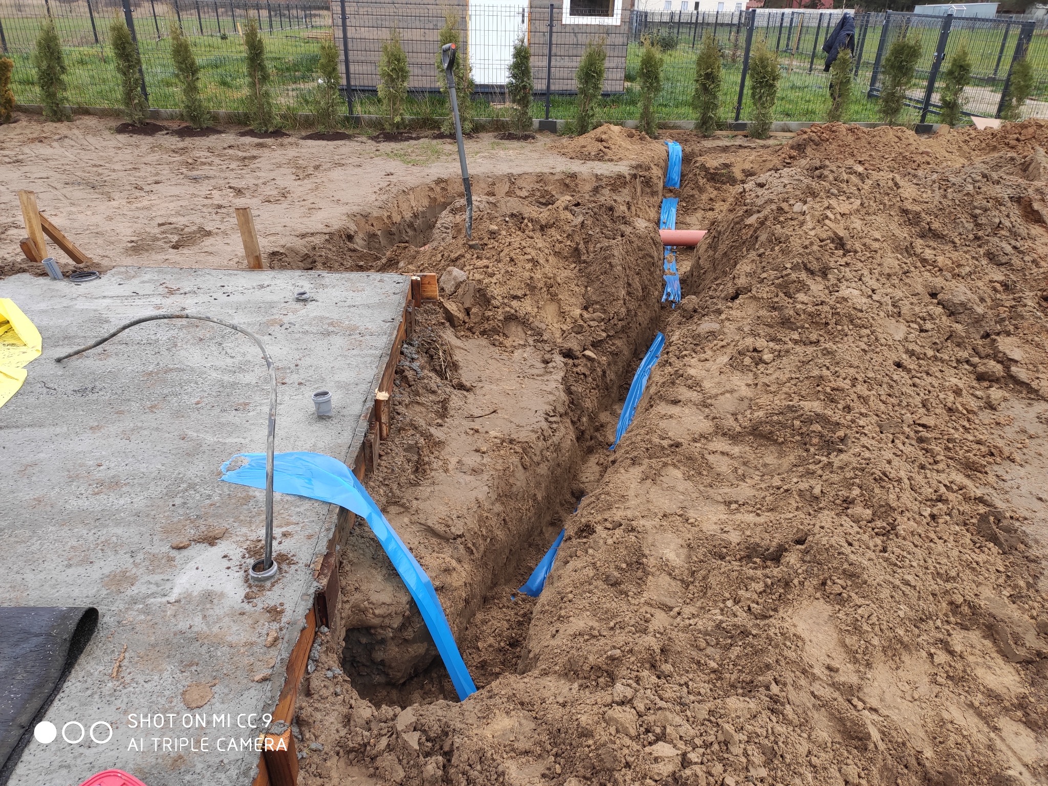 Wykop w ziemi z widocznymi niebieskimi rurami osłonowymi i rurą w kolorze terakoty, prawdopodobnie instalacja pod ogrzewanie podłogowe, widoczny betonowy fundament budynku, szpadel wbity w ziemię...