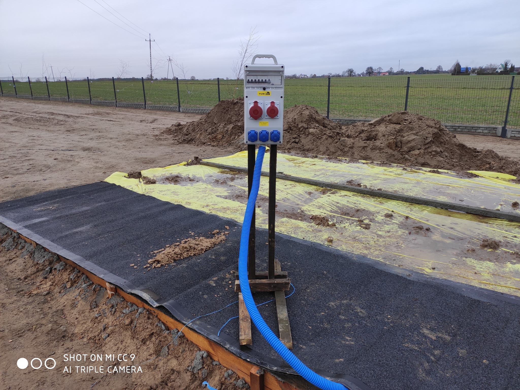 Tymczasowa skrzynka elektryczna z gniazdami siłowymi, zamontowana na drewnianej konstrukcji na placu budowy, z niebieskim karbowanym przewodem. W tle pryzmy ziemi i ogrodzenie.