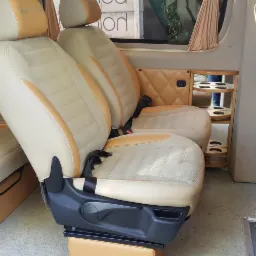 Wnętrze luksusowego busa z obrotowymi fotelami w beżowo-brązowej kolorystyce, z widocznym uchwytem na napoje w tylnej części.