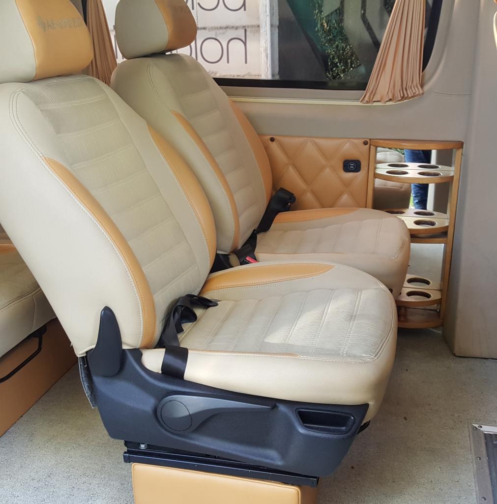 Wnętrze luksusowego busa z obrotowymi fotelami w beżowo-brązowej kolorystyce, z widocznym uchwytem na napoje w tylnej części.