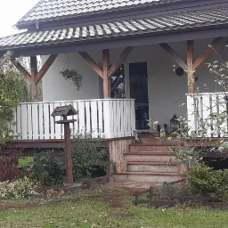 Zadaszony taras domu z drewnianymi słupami i białą balustradą, widok z ogrodu z karmnikiem dla ptaków.