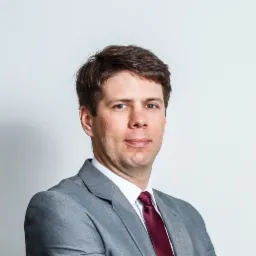 Portret mężczyzny w szarym garniturze i bordowym krawacie na jasnym tle.