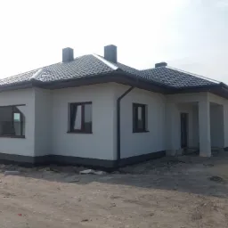 Nowo wybudowany, parterowy dom z szarą elewacją, ciemną dachówką i trzema kominami, widoczny z zewnątrz na tle nieba.