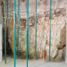 Turkusowe stemple budowlane podtrzymujące drewniany strop w surowym wnętrzu z kamienną ścianą, widoczna rura wentylacyjna i logo firmy MAXWER.