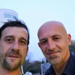 Selfie dwóch mężczyzn, jeden w kamizelce odblaskowej, na tle placu budowy z widocznym zbiornikiem i konstrukcją szkieletową.