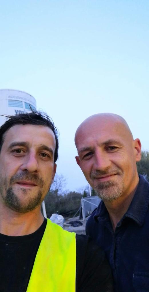 Selfie dwóch mężczyzn, jeden w kamizelce odblaskowej, na tle placu budowy z widocznym zbiornikiem i konstrukcją szkieletową.