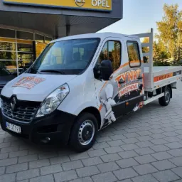Biały samochód dostawczy Renault Master z grafiką przedstawiającą prace tynkarskie, zaparkowany przed salonem Opla na brukowanej nawierzchni.