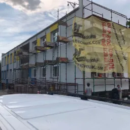 Budynek w trakcie ocieplania elewacji, widoczne rusztowania, siatka ochronna z reklamą firmy budowlanej i pracownicy zbierający odpady budowlane, fragment dachu białego samochodu w dolnej części...