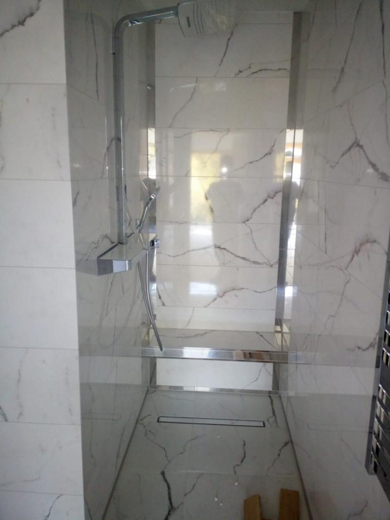 Nowoczesny prysznic typu walk-in z białymi płytkami imitującymi marmur i chromowanymi wykończeniami, widoczny odpływ liniowy.