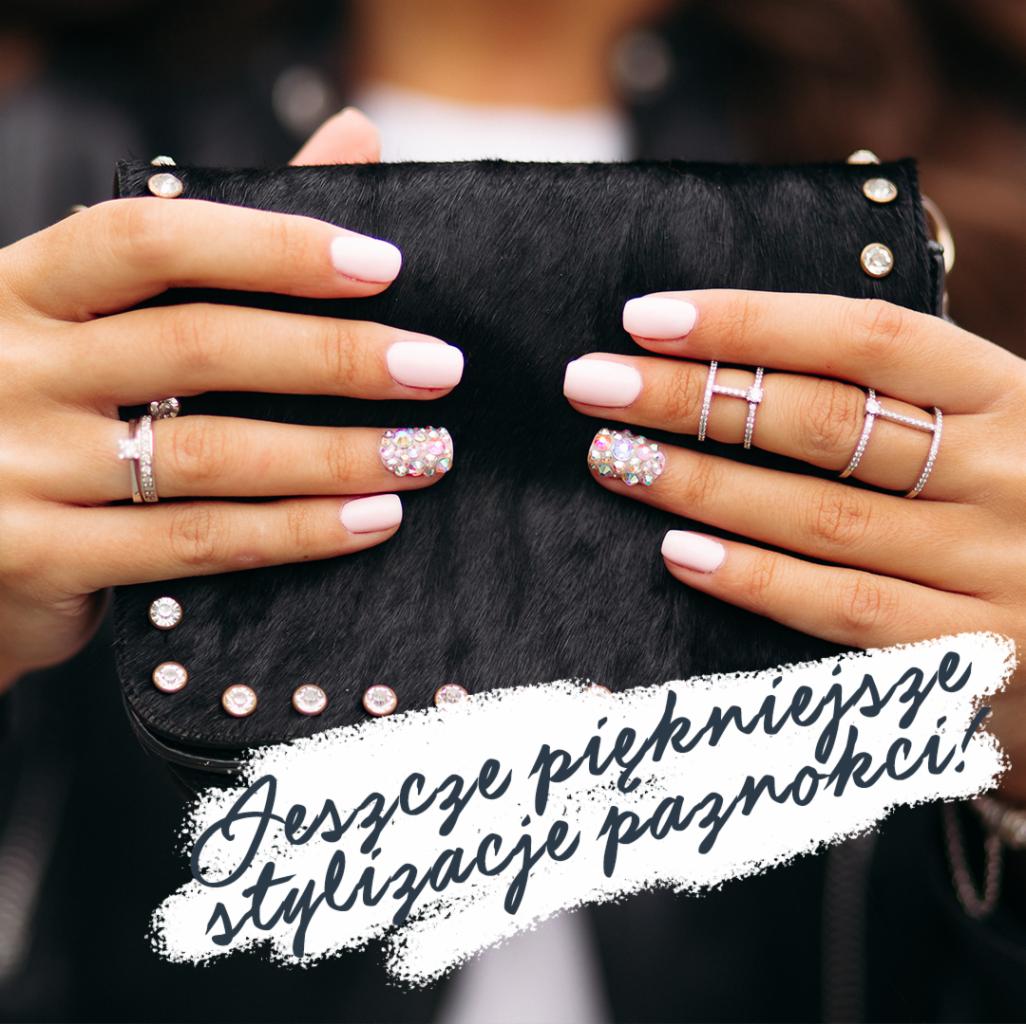 Kobiecie dłonie z jasnoróżowym manicurem, zdobione cyrkoniami na dwóch paznokciach, pierścionki na palcach, trzymające czarną torebkę z ćwiekami.