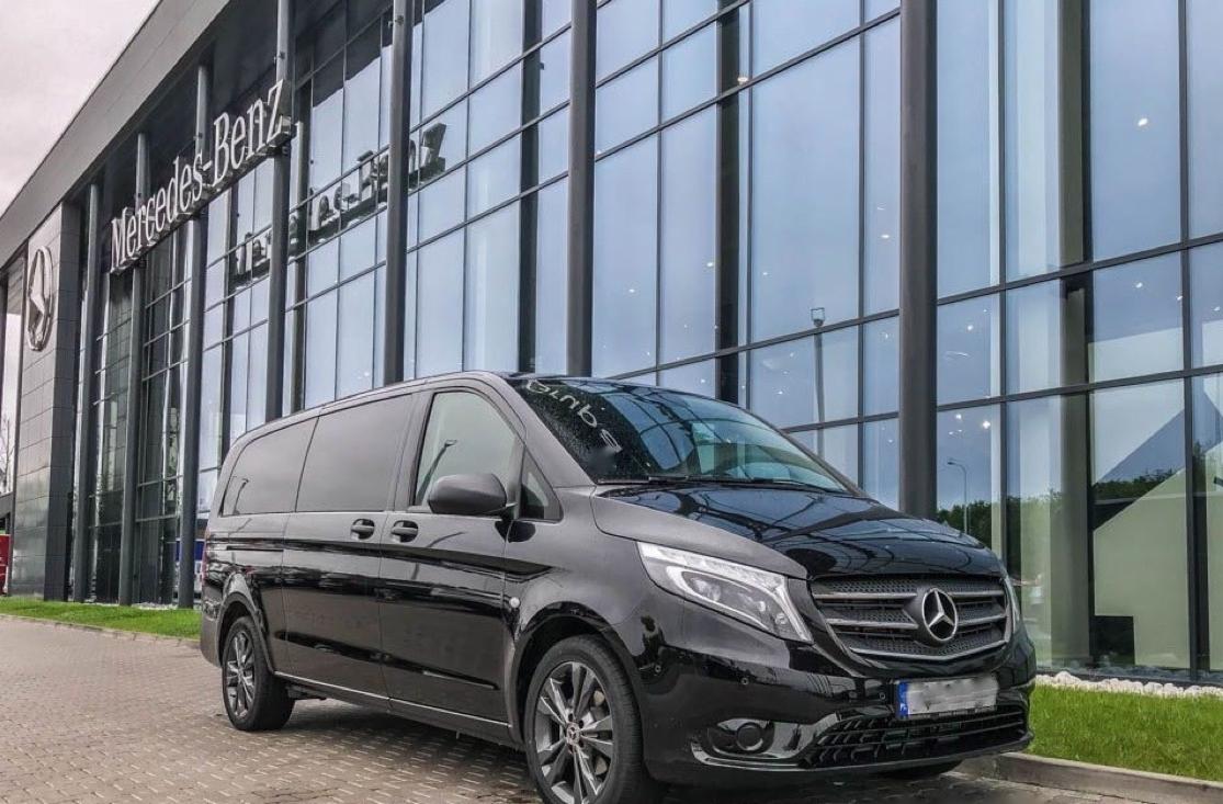 Czarny van Mercedes-Benz V-klasy zaparkowany przed nowoczesnym budynkiem z przeszkloną fasadą i logo Mercedes-Benz.