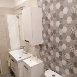 Wykończona łazienka z heksagonalnymi płytkami w odcieniach szarości i bieli, białą armaturą, szafkami pod umywalką i wiszącą, oraz szarą podłogą. Na umywalce stoi mydło w płynie.