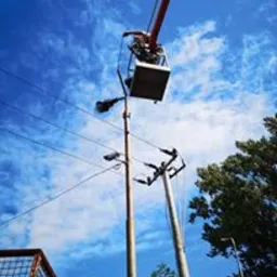 Pracownik na podnośniku koszowym, pracujący przy linii wysokiego napięcia na tle błękitnego nieba z chmurami. Widoczne słupy elektryczne i przewody.