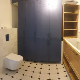 Nowoczesna łazienka w trakcie wykończenia: podwieszana toaleta, szafki w kolorze granatowym, drewniana zabudowa wanny z otwartymi półkami, płytki podłogowe w biało-czarną szachownicę.