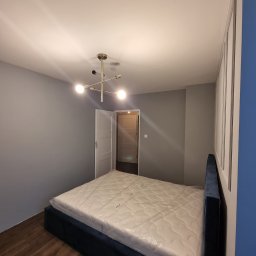 A&A NEW HOME Sp. z o.o. - Nowe łóżko z granatowym, welurowym obiciem i materacem w folii ochronnej w pokoju z szarymi ścianami i drewnianą podłogą. W tle widoczne białe drzwi.