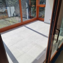 A&A NEW HOME Sp. z o.o. - Jasne płytki podłogowe imitujące kamień, ułożone w przeszklonym pokoju z drewnianymi oknami, widok na zewnątrz z drzewami i niebieskim niebem, cień rzucany przez otwarte drzwi balkonowe.