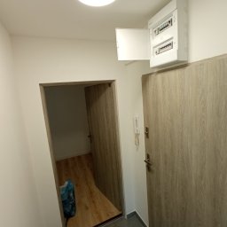 A&A NEW HOME Sp. z o.o. - Wnętrze mieszkania po remoncie: widoczna skrzynka elektryczna z otwartymi drzwiczkami nad drzwiami wejściowymi, obok drugie drzwi do pokoju z panelami na podłodze, w rogu niebieski worek na śmieci.