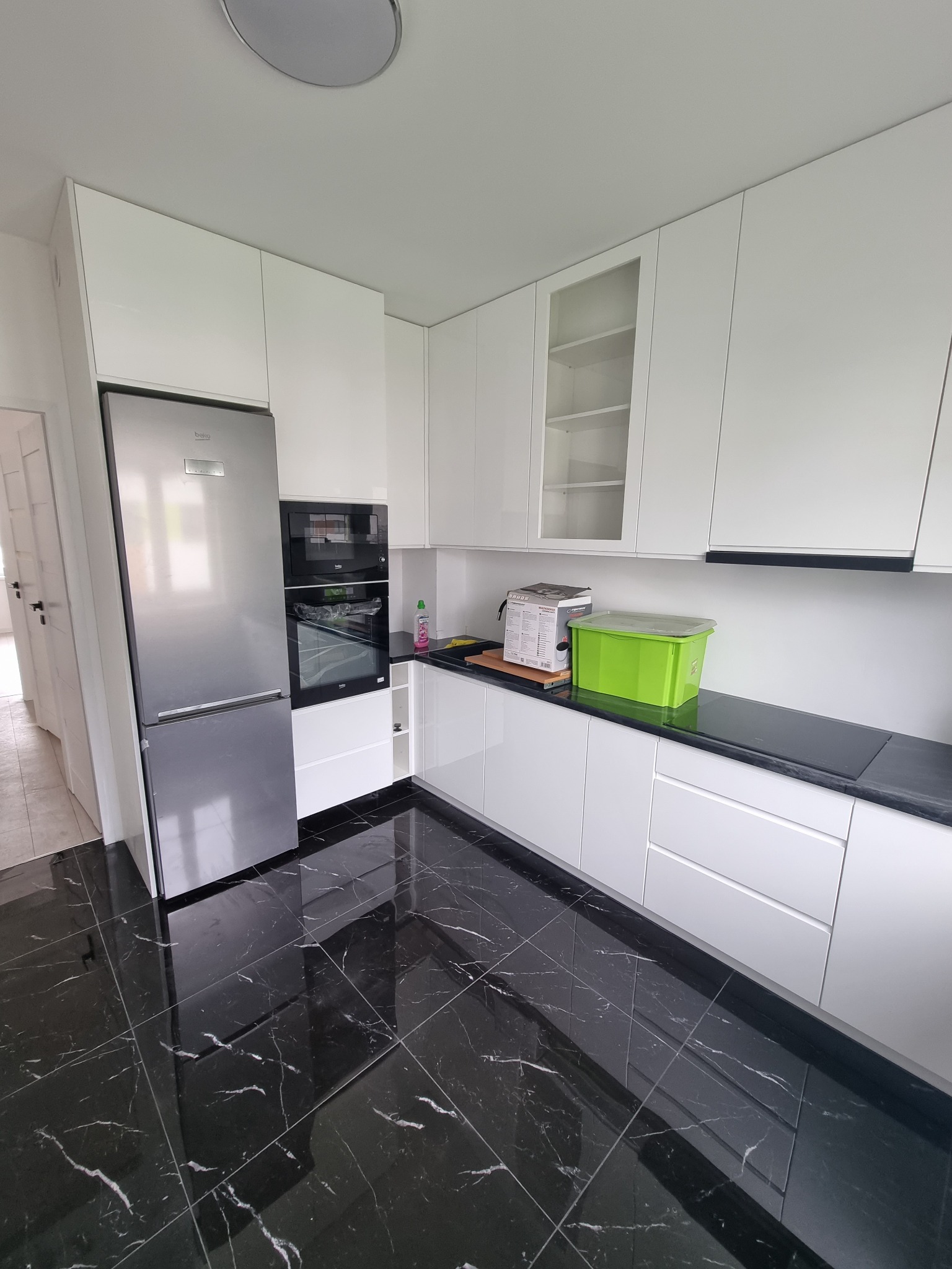 Nowoczesna kuchnia z błyszczącymi, czarnymi płytkami podłogowymi imitującymi marmur, białe szafki z frontami na wysoki połysk, srebrna lodówka Beko i zabudowany piekarnik z mikrofalówką.