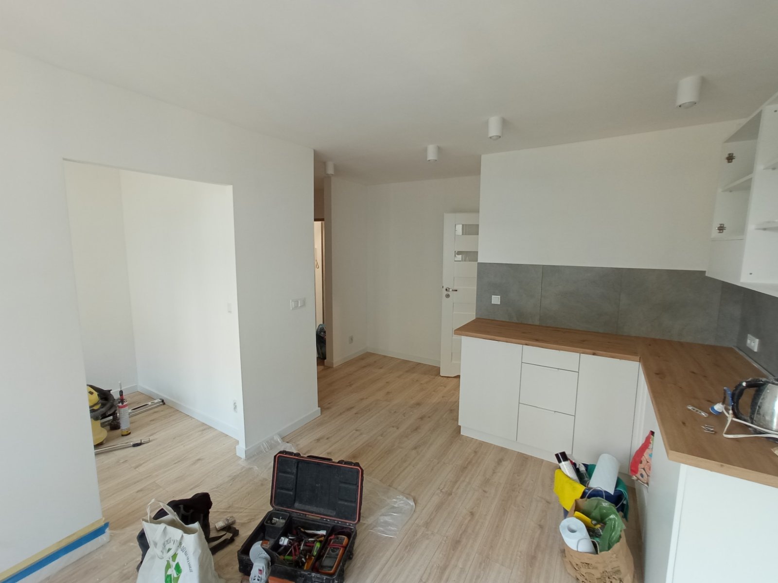 Wnętrze mieszkania w trakcie prac wykończeniowych: widoczna nowa, biała zabudowa kuchenna z drewnianym blatem i szarą płytką na ścianie, parkiet na podłodze, narzędzia i akcesoria malarskie w rogu...