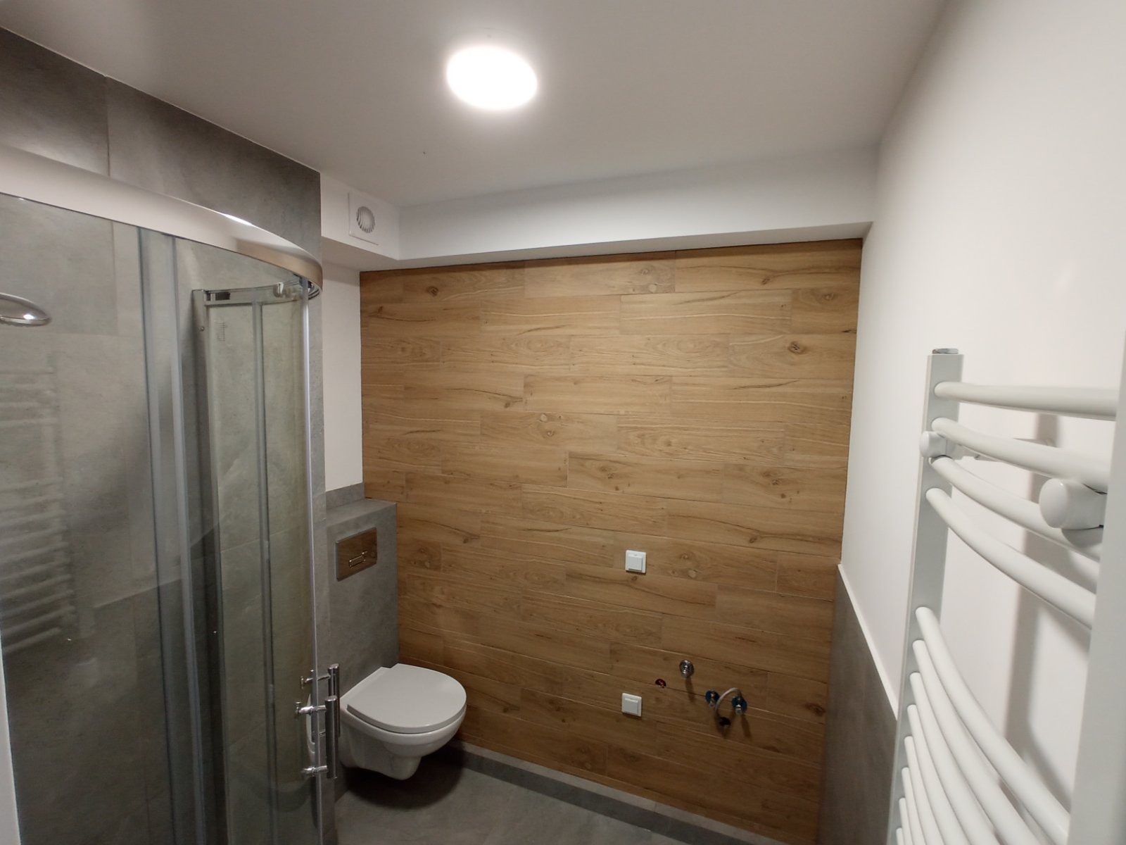 Nowoczesna łazienka z kabiną prysznicową, toaletą podwieszaną i grzejnikiem drabinkowym na tle drewnopodobnej ściany, widoczne przyłącza wodne i elektryczne.