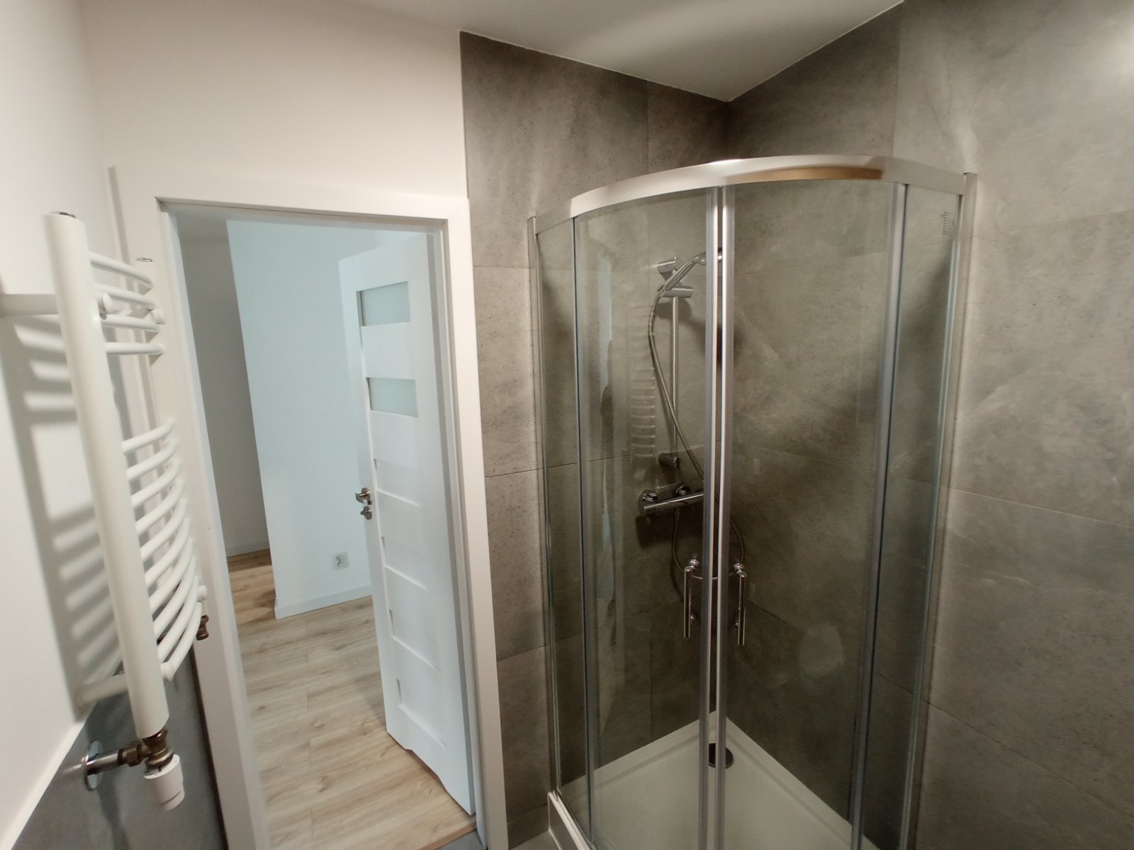 Nowoczesna łazienka z kabiną prysznicową typu walk-in, szare płytki imitujące beton, biały grzejnik drabinkowy i częściowo otwarta biała stolarka drzwiowa z poziomymi przeszkleniami.