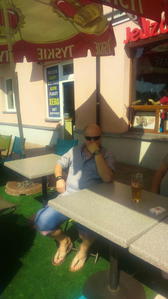 Mężczyzna w okularach siedzi przy stoliku na zewnątrz lokalu z szyldem reklamowym nadruku i reklamy.