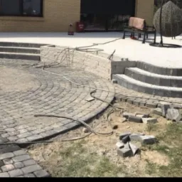 Układanie kostki brukowej na tarasie z kilkoma poziomami schodów, widoczne niedokończone fragmenty i porozrzucane kostki, w tle budynek mieszkalny i elementy wyposażenia ogrodu.