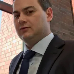 Portret mężczyzny w garniturze, stojącego na tle ceglanej ściany, z widocznym fragmentem okna w tle.