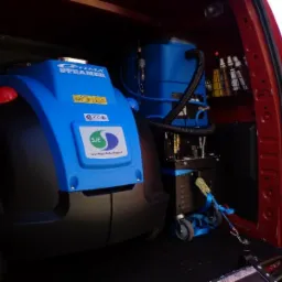 Wnętrze czerwonego samochodu dostawczego z profesjonalnym, niebieskim sprzętem do czyszczenia tapicerki, w tym Optima Steamer i dodatkowe butelki z detergentami na półce.