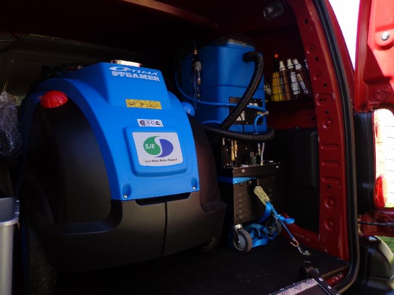 Wnętrze czerwonego samochodu dostawczego z profesjonalnym, niebieskim sprzętem do czyszczenia tapicerki, w tym Optima Steamer i dodatkowe butelki z detergentami na półce.