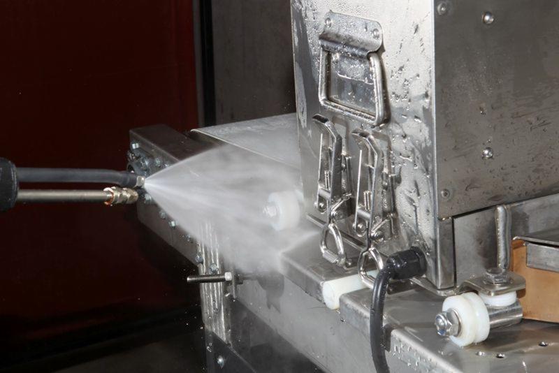 Strumień wody pod ciśnieniem z dyszy czyszczący metalową konstrukcję z uchwytami i kółkami, widoczne ślady wilgoci.