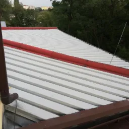 Biała blacha trapezowa na dachu z czerwonymi elementami wykończeniowymi, widoczny brązowy rzygacz, w tle zieleń drzew i zabudowa miejska.