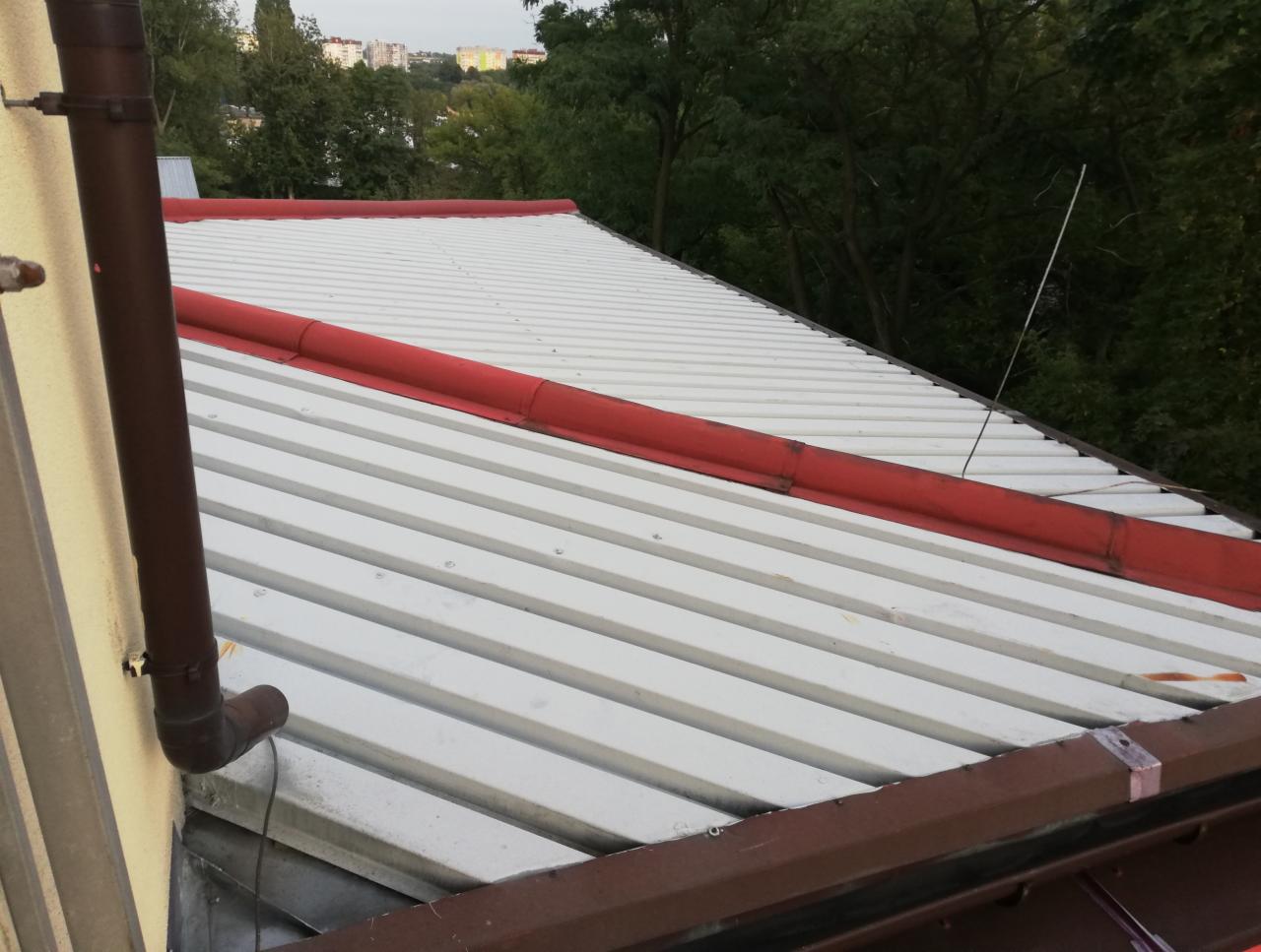 Biała blacha trapezowa na dachu z czerwonymi elementami wykończeniowymi, widoczny brązowy rzygacz, w tle zieleń drzew i zabudowa miejska.