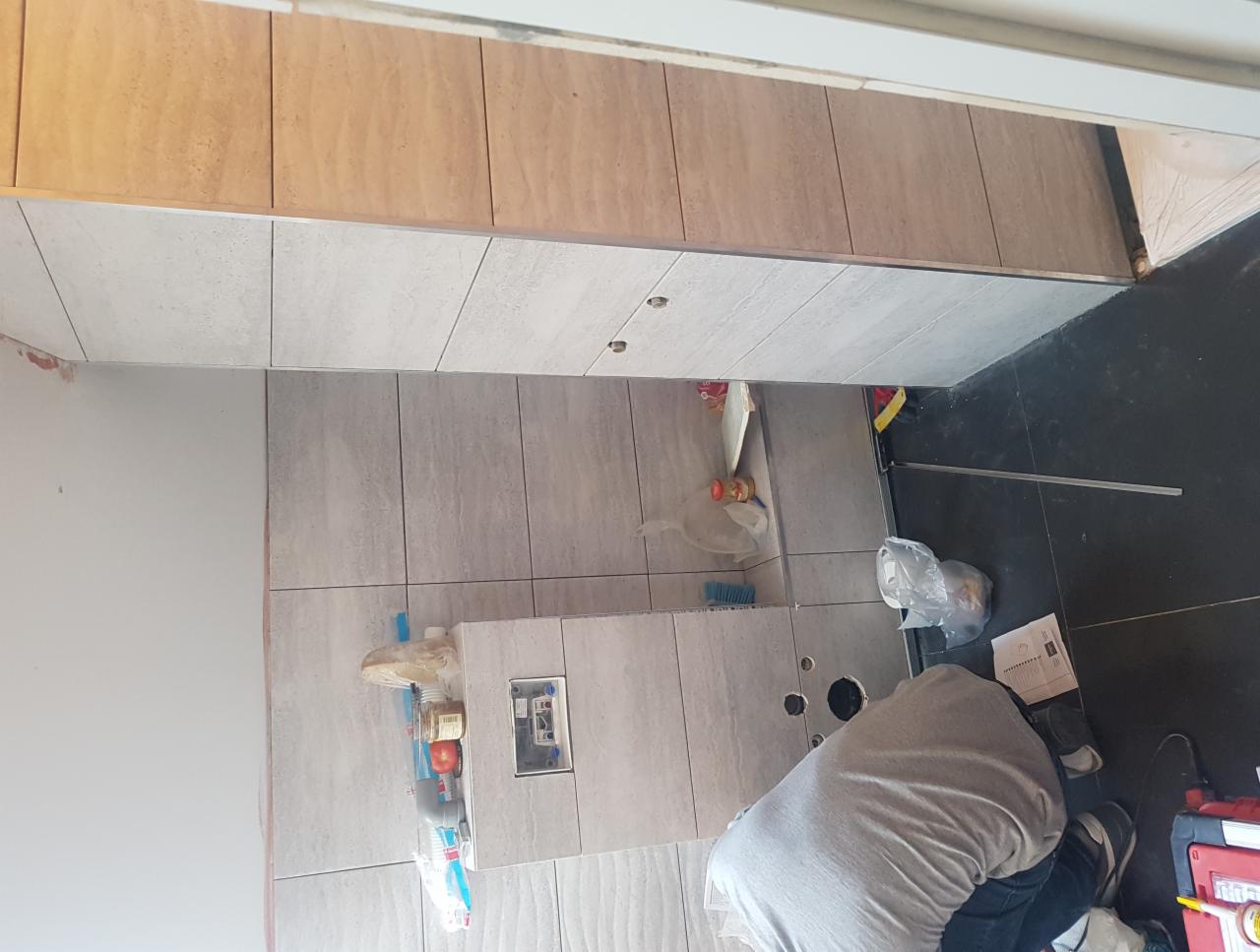 Wnętrze łazienki w trakcie prac. Widoczne ułożone jasne płytki, instalacja pod umywalkę i pracownik w trakcie montażu. Ujęcie z góry, z widocznymi narzędziami.